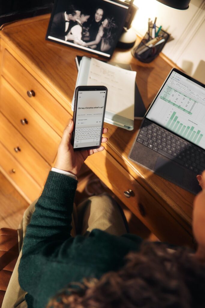 Person holding phone near laptop and tablet on desk